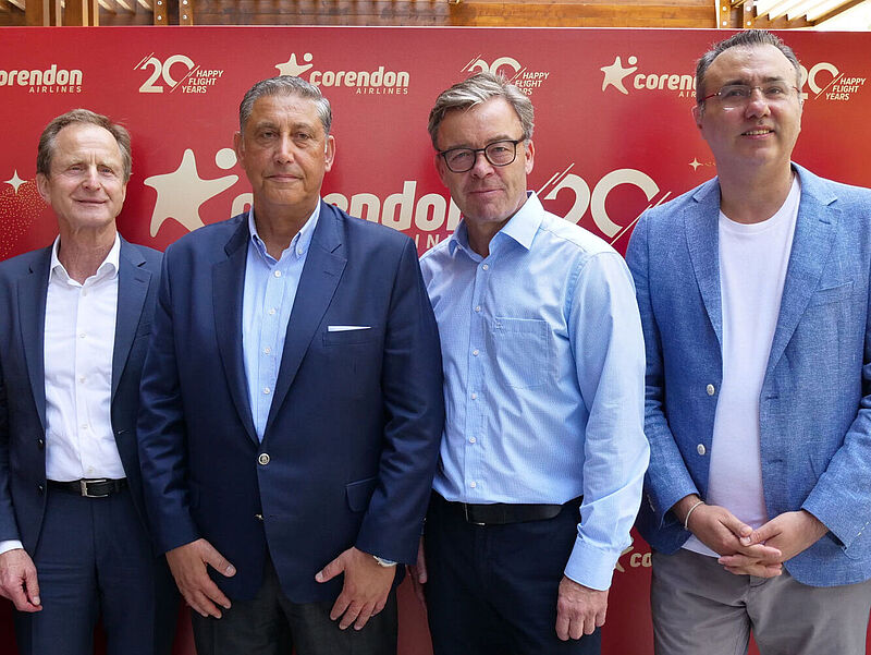 Vier Manager in legerer Business-Kleidung stehen nebeneinander und lächeln in die Kamera. Im Hintergrund steht eine rote Stellwand mit Logos der Fluglinie Corendon Airlines.