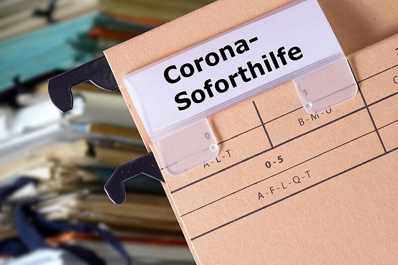 Eine braune Hängeregistraturmappe mit dem beschrifteten Reiter „Corona-Soforthilfe“ ist im Vordergrund zu sehen. Im Hintergrund befinden sich weitere Aktenmappen in verschiedenen Farben, die unscharf dargestellt sind. Die Szene deutet auf Verwaltung oder Archivierung von Anträgen zur Corona-Soforthilfe hin.