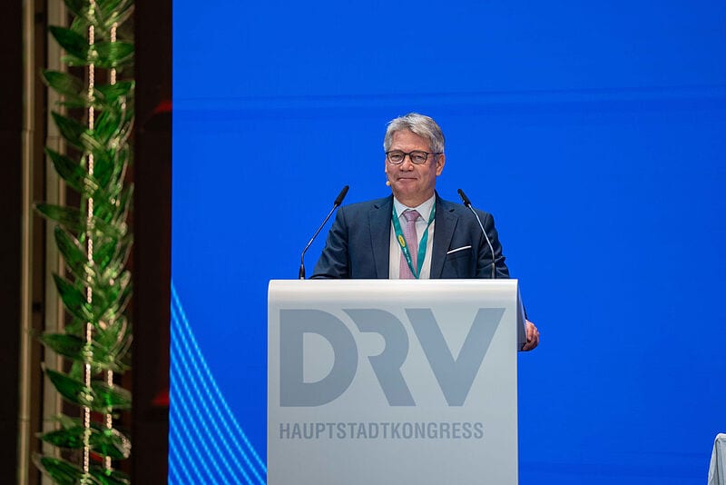 Ein Manager mittleren Alters in einem grauen Anzug steht lächelnd auf einem Rednerpult mit der Aufschrift DRV vor einer blauen Leinwand