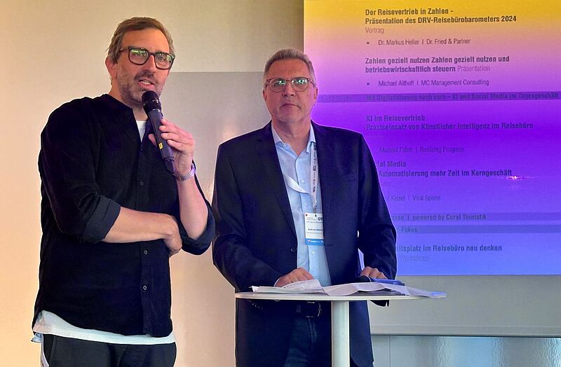 Zwei Männer stehen nebeneinander auf einer Bühne. Der Mann links spricht in ein Mikrofon, der rechte steht hinter einem Stehtisch mit Papieren. Im Hintergrund ist ein farbig beleuchteter Bildschirm mit dem Veranstaltungsprogramm des DRV-Events „Tag des Reisevertriebs 2024“ zu sehen.