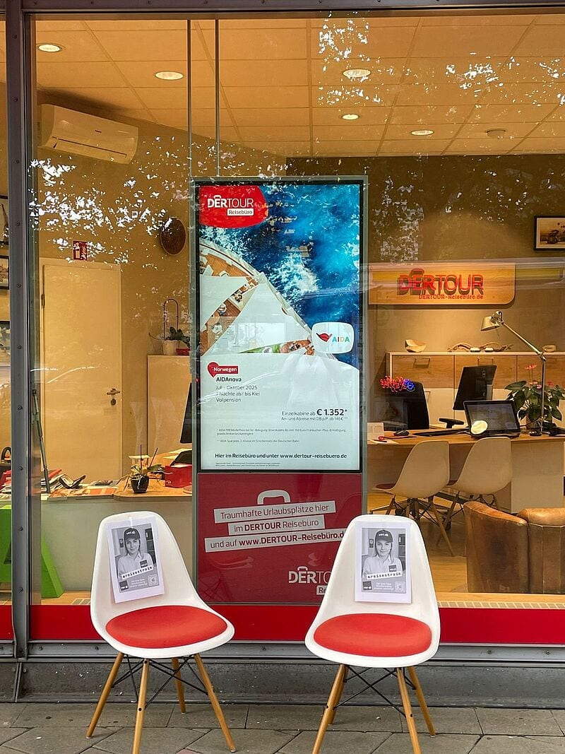 Zwei leere Stühle mit ver.di-Streikplakaten stehen vor einem Dertour-Reisebüro. Im Hintergrund ist durch die große Fensterscheibe der modern eingerichtete Innenraum des Büros sichtbar, inklusive Schreibtischen, Werbematerial und einem großen Plakat für Aida-Kreuzfahrten. Aufschrift an der Scheibe: „Traumhafte Urlaubsplätze hier im Dertour Reisebüro“. Die Szene signalisiert den laufenden Streik durch symbolische Abwesenheit der Mitarbeitenden. 
