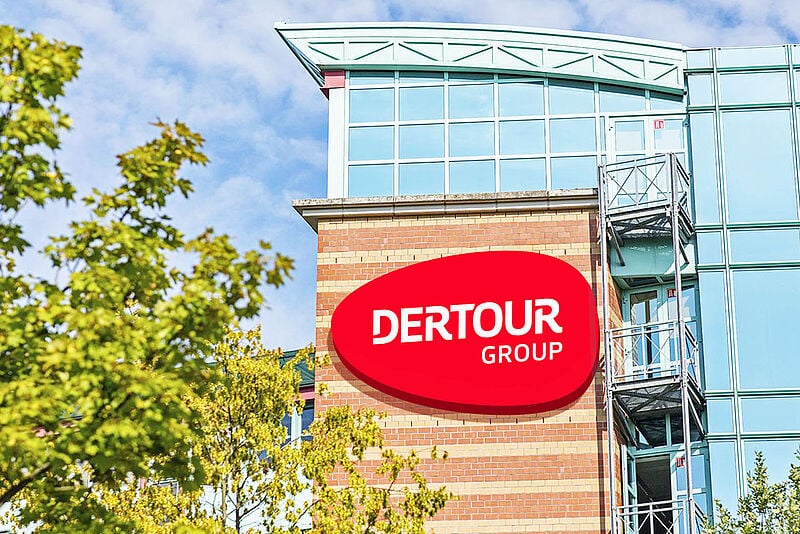 Außenansicht eines modernen Bürogebäudes mit rotem Dertour Group-Logo auf einer Backsteinfassade. Im Vordergrund sind grüne Bäume und ein blauer Himmel mit vereinzelten Wolken zu sehen.