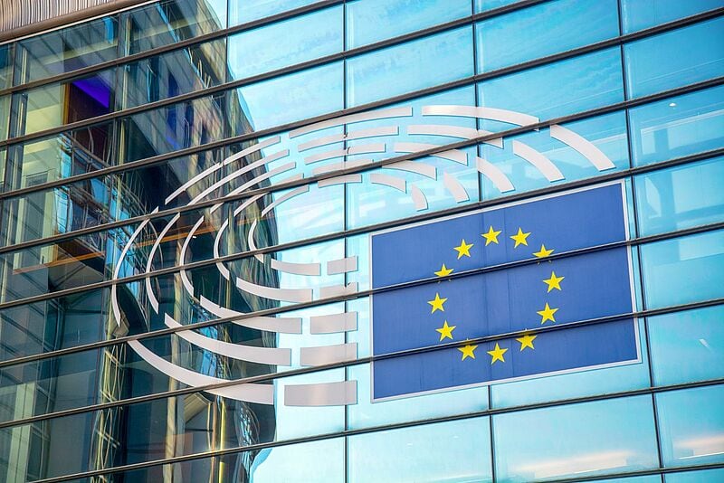 Fassade des Europäischen Parlaments in Brüssel mit dem Emblem des Parlaments und der EU-Flagge – zu sehen sind reflektierende Glasfenster, in denen sich ein gegenüberliegendes Gebäude spiegelt.