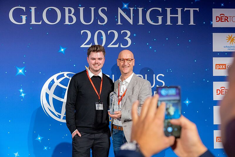 Zwei Männer stehen lächelnd vor einer blauen Wand mit der Aufschrift „GLOBUS NIGHT 2023“, einer davon hält einen Glaspokal in der Hand. Beide tragen rote Veranstaltungsausweise um den Hals. Im Vordergrund wird die Szene von einer Person mit Smartphone fotografiert.