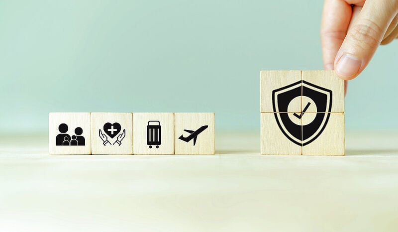 Holzwürfel mit Reiseversicherungs-Icons: Personen, Gesundheit, Gepäck, Flugzeug und Schutzschild-Symbol, auf mintgrünem Hintergrund.