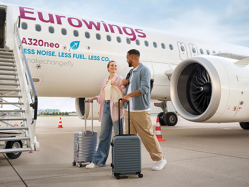 Ein junges Paar geht mit Rollkoffern eines Eurowings-Flugzeugs entlang hin zur Einstiegstreppe, um an Bord zu gehen.