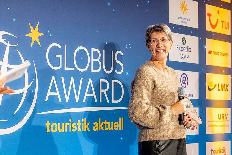 Verleihung des Globus Award von touristik aktuell – Gewinnerin mit kurzen Haaren und mit braunem Pullover auf der Bühne mit Auszeichnung und Mikrofon beim Branchenevent der Touristik“