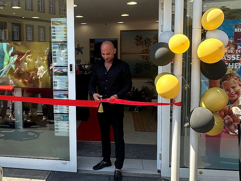 Ein Mann in schwarzer Kleidung durchschneidet mit einer Schere ein rotes Band bei der Eröffnung eines Geschäfts. Er steht vor einem modernen Eingangsbereich mit Glastüren, an denen schwarze, gelbe und graue Luftballons befestigt sind. Im Inneren ist ein roter Teppich ausgerollt, und die Wände sind mit Urlaubsbildern dekoriert. In der Spiegelung der Glasscheibe sind mehrere Personen zu sehen, die Fotos machen.