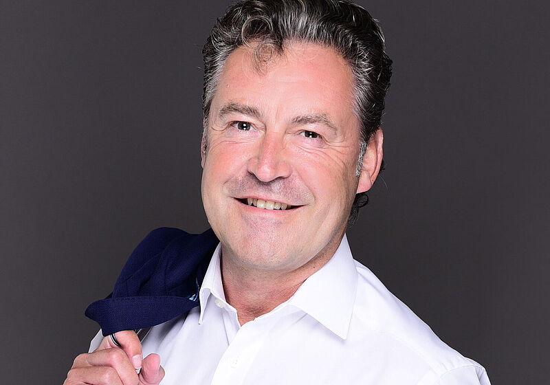 Ein Mann mittleren Alters mit dunkelgrauem Haar posiert lächelnd für ein Porträtfoto. Er trägt ein weißes Oberhemd und hält sein Sakko lässig über die Schulter.