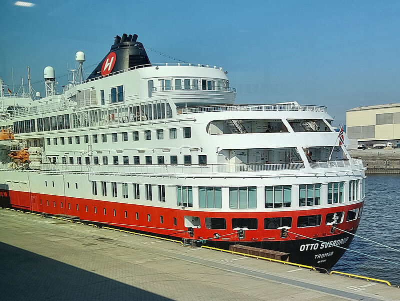 Ein kleines Kreuzfahrtschiff mit rot-schwarzem Rumpf und weißem Aufbau liegt vertäut an einer Pier. Am Heck prangt in weißen Lettern der Schiffsname Otto Sverdrup. Die Sonne scheint.