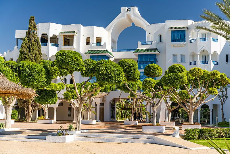 Hinter grünen Bäumen steht ein weißes Hotelgebäude