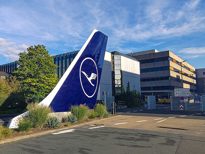 Großes blaues Lufthansa-Leitwerk mit Kranich-Logo vor modernem Bürogebäude am Frankfurter Flughafen, umgeben von Grünanlagen und Parkplätzen.