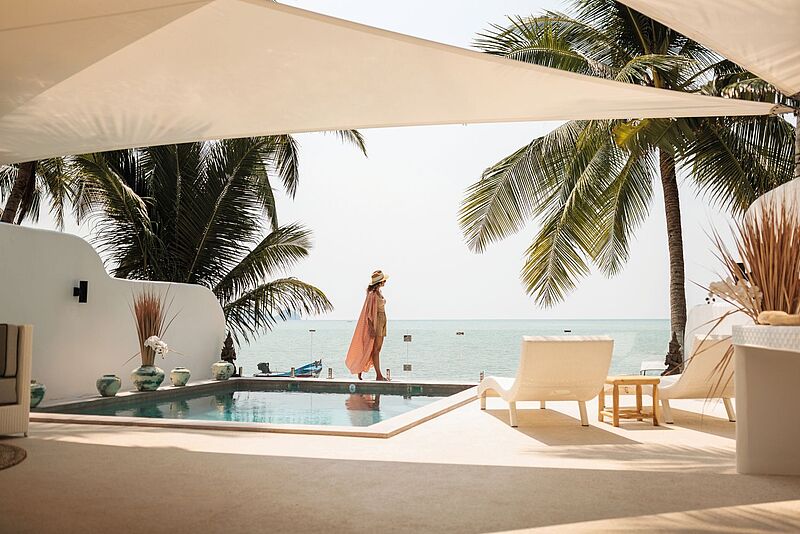 Frau in Sommerkleid und Hut steht am Rand eines Infinity-Pools mit Blick auf das Meer, umgeben von Palmen und moderner, offener Architektur in einem luxuriösen Strandresort.