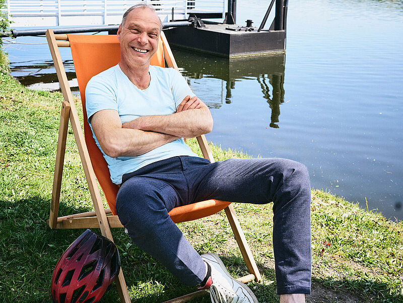 Der Schauspieler Martin Brambach, mit schütterem Haar und strahlendem Lächeln, sitzt in Freizeitkleidung in einem Liegestuhl an einem See und schaut in die Kamera. Die Sonne scheint.