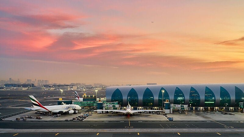 Das moderne Terminal des Flughafens Dubai bei Sonnenuntergang mit türkis verglasten Bögen, Emirates-Flugzeugen am Gate und einem dramatischen rosa-goldenen Himmel.