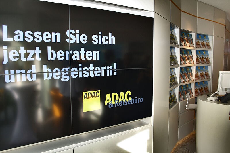 Ein modernes ADAC-Reisebüro mit einem großen digitalen Bildschirm auf der linken Seite, der die Botschaft „Lassen Sie sich jetzt beraten und begeistern!“ in weißer Schrift auf schwarzem Hintergrund zeigt. Darunter sind die Logos des ADAC und des ADAC Reisebüros zu sehen. Rechts im Bild befinden sich weiße Regale mit Reisebroschüren, die ordentlich in Reihen angeordnet sind. Im Vordergrund steht ein geschwungener weißer Beratungstresen mit einem Computerbildschirm. Die Atmosphäre ist hell und einladend.
