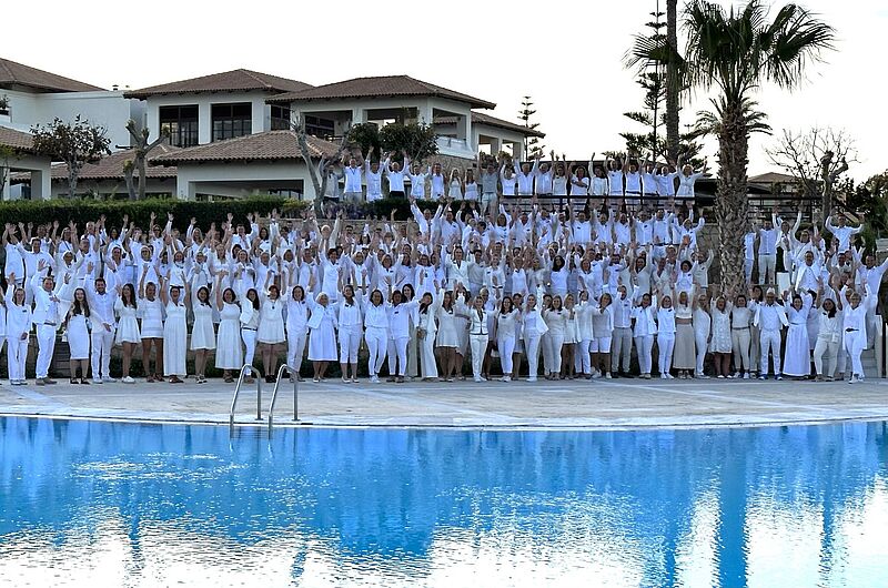 Gruppenfoto mit einer großen Anzahl von Menschen, die alle in weißer Kleidung gekleidet sind. Sie stehen in mehreren Reihen aufgestaffelt vor einem Hotel mit mediterraner Architektur. Viele heben die Arme freudig in die Luft. Im Vordergrund befindet sich ein Swimmingpool, der das Bild und die Personen leicht spiegelt. Palmen und gepflegte Gärten rahmen die Szene.