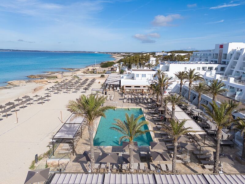 Ein ansprechender weißer Hotelbau an einem hellen feinsandigen Strand mit Liegen, dazwischen ein Pool.