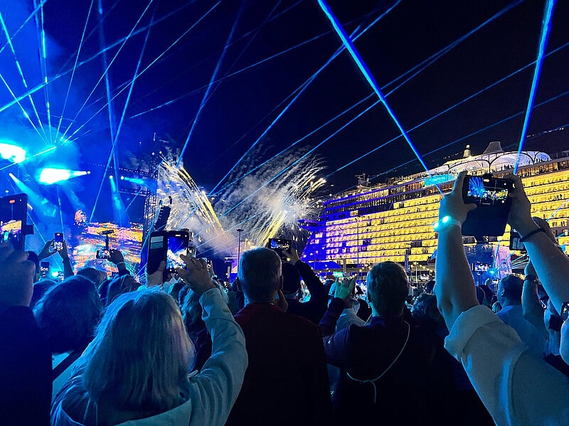 Eine große Menschenmenge steht bei Nacht vor einer Bühne und einem beleuchteten Kreuzfahrtschiff. Viele Personen halten ihre Handys hoch