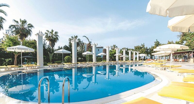 Sonniger Außenbereich eines Hotels mit einem großen, runden Swimmingpool, umgeben von gelben Liegestühlen und weißen Sonnenschirmen, gesäumt von Palmen und dekorativen Säulen im klassischen Stil