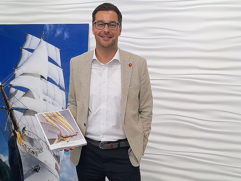 Ein lächelnder Reederei-Manager in den Mittvierzigern mit braunem Haar und Brille steht vor einem Roll-up mit einem weißen Segelkreuzfahrtschiff. Er trägt ein beiges Sakko, ein weißes Hemd und Jeand und hält in der Hand einen Kreuzfahrtkatalog.
