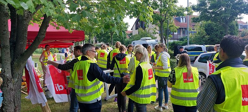 Gruppe von Verdi-Mitgliedern in gelben Warnwesten bei einer Streikversammlung im Freien unter Bäumen. Die Personen stehen in kleinen Gruppen zusammen, einige halten Fahnen oder sprechen miteinander. Im Hintergrund sind rote Pavillons, Wohnhäuser und ein Polizeifahrzeug zu sehen.