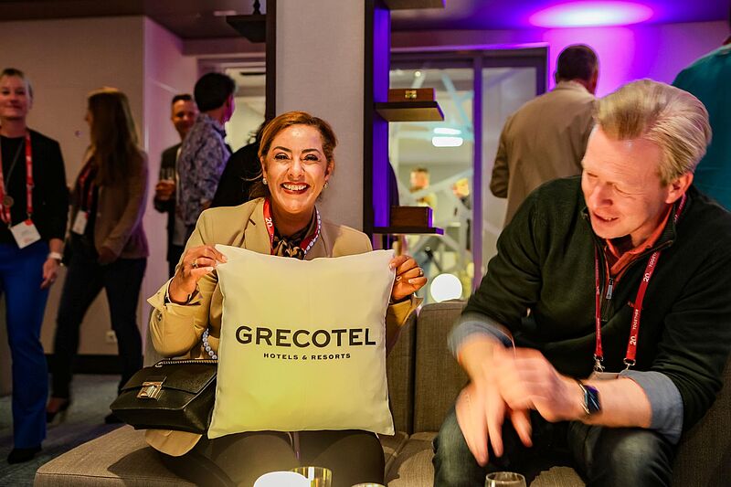 Ein Frau und ein Mann sitzen in einer Bar. Die Frau hält ein Kissen in der Hand mit der Aufschrift Grecotel.