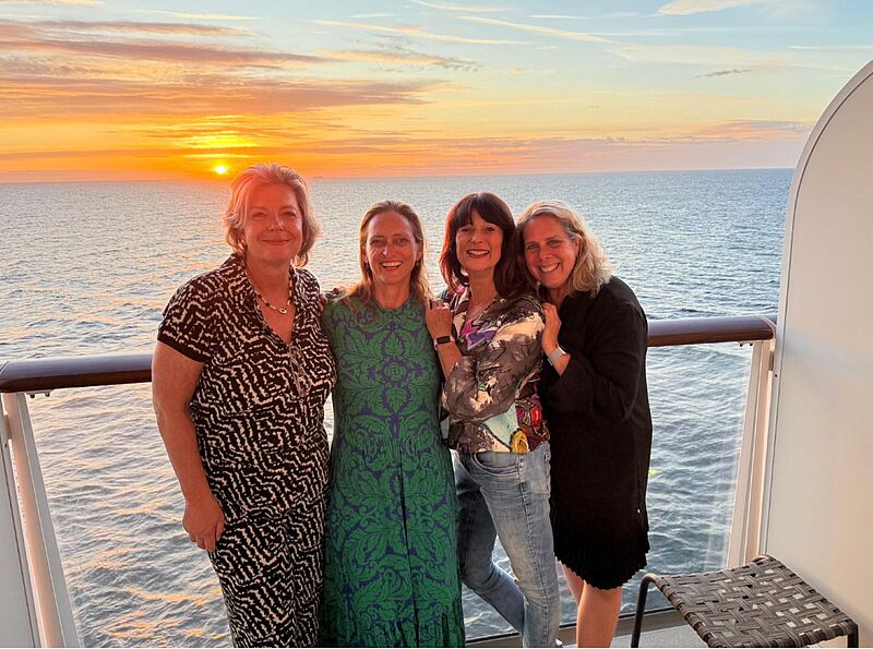 Vier Frauen in Abendgarderobe auf einem Balkon. Im Meer geht die Sonne unter