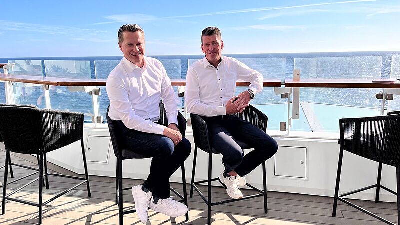 Zwei Männer mit dunklen Hosen und weißem Hemd sitzen auf einem Stuhl und lächeln in die Kamera. Hinter ihnen das blaue Meer. Die Sonne scheint