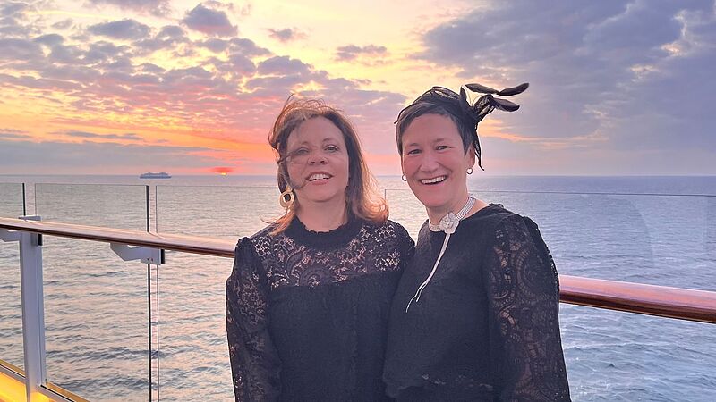 Zwei elegant gekleidete Frauen lehnen an der Reling. Der Wind zerzaust die Haare