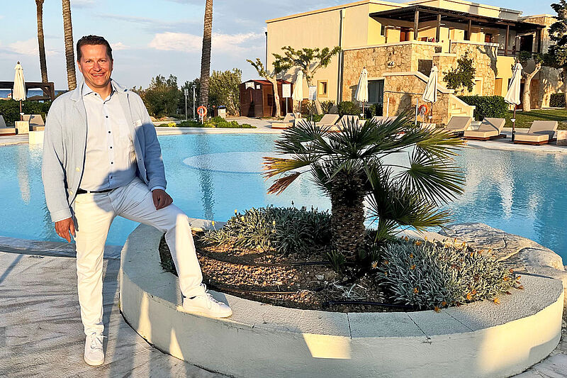 Ein lächelnder Mann, es ist Benjamin Jacobi von TUI Deutschland, steht am Rand eines eleganten Pools im Grecotel Imperial auf Kos. Er trägt ein hellblaues Sakko, ein helles Hemd, weiße Hose und weiße Sneaker. Eine seiner Füße ruht auf dem erhöhten, runden Pflanzbeet mit einer kleinen Palme und Bodendeckern. Im Hintergrund sind Sonnenliegen, Sonnenschirme, Palmen und ein modernes Gebäude mit Natursteinfassade und Terrasse zu sehen. Die Sonne wirft lange Schatten, was auf den späten Nachmittag hinweist.