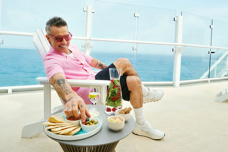 Mann in rosa Hemd entspannt auf Kreuzfahrtschiff-Balkon, genießt Snacks und Getränke mit Meerblick.