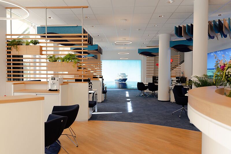 Innenansicht des neuen TUI-Flagshipstores in Aschaffenburg: moderne Beratungslounge mit hellen Holzelementen, blauen Stoffdecken im Wellen-Design, Pflanzen und offenen Beratungsplätzen. Im Hintergrund ein großes Bildschirmbild mit einem Kreuzfahrtschiff.
