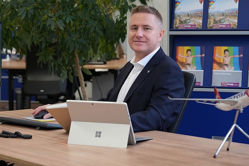 Geschäftsmann im Anzug an einem Schreibtisch mit katalogwand im Hintergrund. Der Mann schaut freundlich in die Kamera.