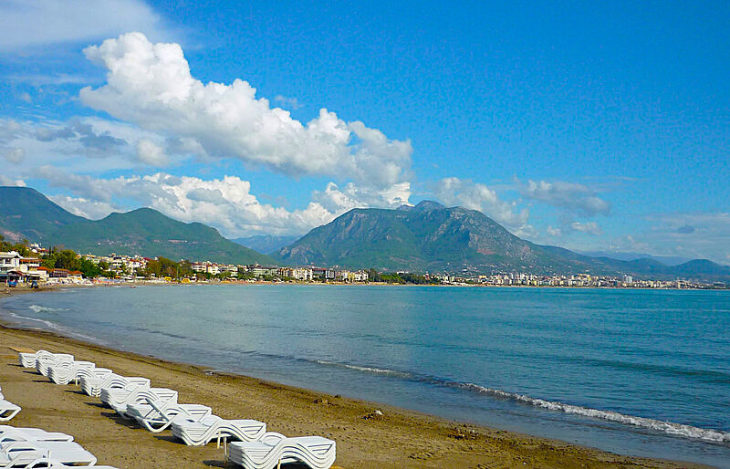 Strand von Alanya in der Türkei mit leeren weißen Sonnenliegen im Vordergrund, ruhigem Meer und einer Stadt entlang der Küste. Im Hintergrund erheben sich grüne Berge unter einem teils bewölkten, blauen Himmel.