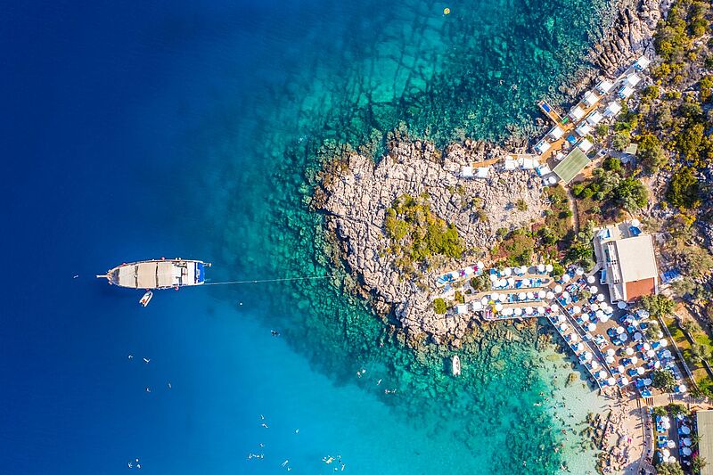 Luftaufnahme eines malerischen Küstenabschnitts mit kristallklarem, türkisfarbenem Wasser in Kaş, Antalya (Türkei). Auf der rechten Seite des Bildes befinden sich Felsen und ein beliebter Strandbereich mit zahlreichen weißen Sonnenschirmen und Liegen, eingebettet in mediterrane Vegetation. Mehrere Holzstege führen ins Wasser. Auf der linken Seite ankert ein großes Ausflugsboot im tiefblauen Meer, mit kleineren Booten und badenden Menschen rundherum.