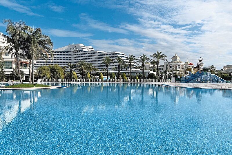 Großer, einladender Hotelpool mit klarem, blauem Wasser vor einer modernen Hotelanlage und Palmen unter blauem Himmel; im Hintergrund ein verspielter Wasserpark und ein prachtvolles Gebäude mit Türmen im orientalischen Stil.