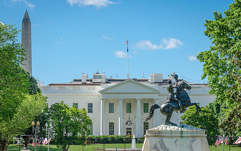 Das Weiße Haus, Sitz des US-Präsidenten, wird von ikonischen Wahrzeichen Washingtons umrahmt. Links im Hintergrund ist das Washington Monument und im Vordergrund eine Reiterstatue umgeben von grünen Bäumen zu sehen.