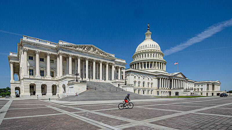 Das Kapitol in der US-Hauptstadt Washington DC vor blauem Himmel an einem sonnigen Sommertag. Der Platz vor dem Gebäude ist leer, lediglich ein Fahrradfahrer fährt durch das Bild.