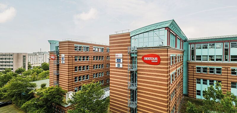 Mehrstöckiges Bürogebäude mit rot-brauner Klinkerfassade und vielen Fenstern, darauf ist ein großes rotes Schild mit der Aufschrift „DERTOUR GROUP“ zu sehen. Das Gebäude liegt inmitten von grünen Bäumen und weiteren modernen Bürohäusern, der Himmel ist leicht bewölkt.