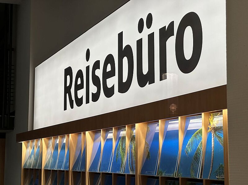 Leuchtendes Schild mit der Aufschrift „Reisebüro“ in schwarzer Schrift auf weißem Hintergrund. Darunter befinden sich zahlreiche Prospektfächer mit Reisebroschüren, die tropische Strandmotive mit Palmen zeigen.