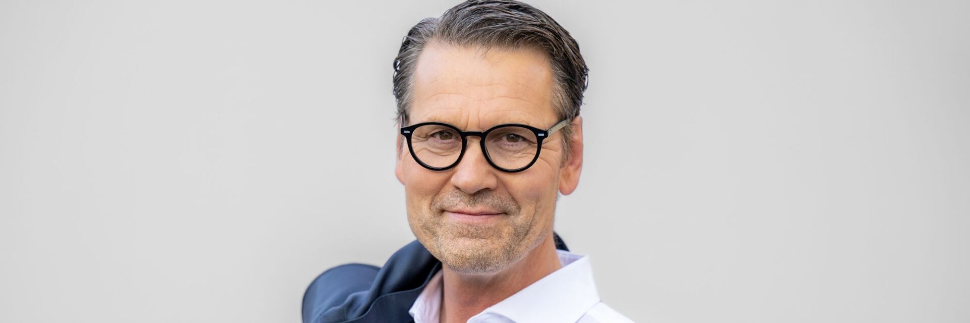 Mann im weißen Hemd hält seine dunkelblaue Anzugjacke lässig über der Schulter, vor einem hellgrauen, einfarbigen Hintergrund.