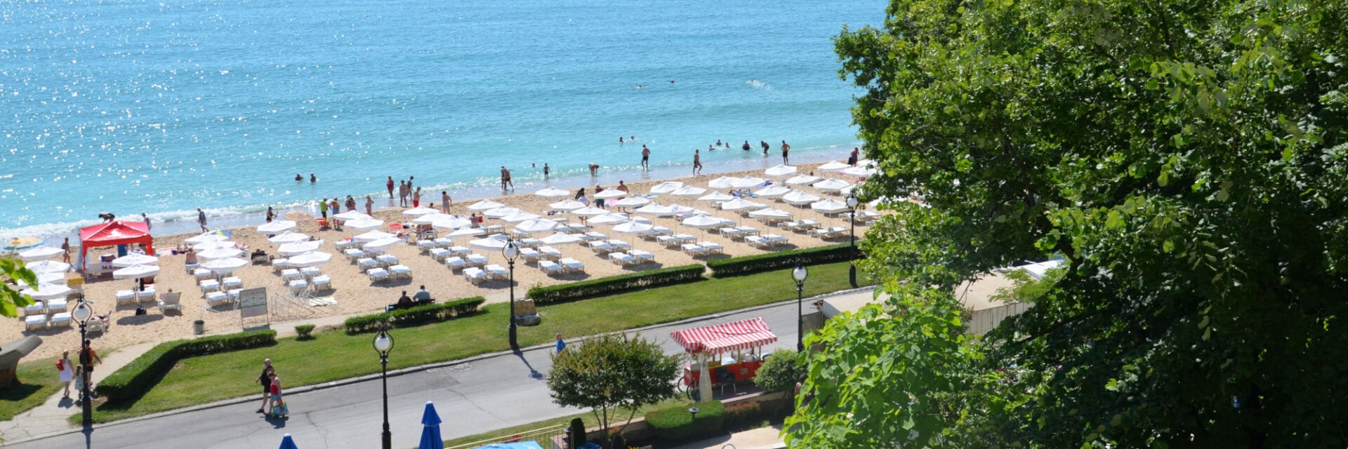 Blick von oben auf einen sonnigen Sandstrand mit vielen weißen Sonnenliegen und Sonnenschirmen, badenden Menschen im türkisblauen Meer und einer Promenade mit Bäumen und kleinen Verkaufsständen im Vordergrund.
