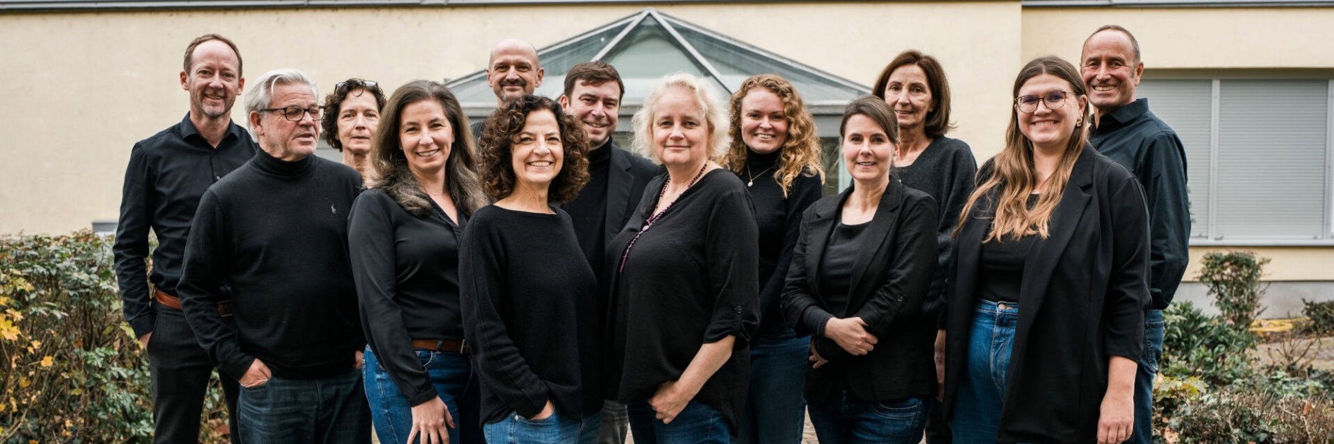 Eine Gruppe von dreizehn Personen steht dicht beisammen im Freien vor einem Gebäude mit einem verglasten Dachaufsatz. Alle tragen dunkle Oberteile und überwiegend Jeans. Die Gruppe lächelt in die Kamera, und im Vordergrund sind Sträucher und herbstliche Bepflanzung zu sehen.