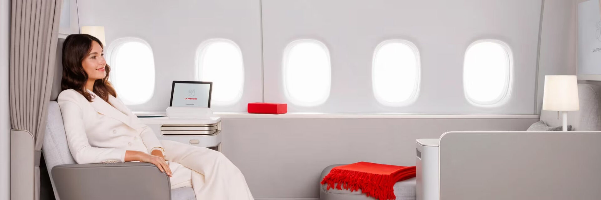 In der luxuriösen First Class einer Airline sitzt eine Frau entspannt in einem großzügigen, hellen Flugzeuginnenraum. Die Kabine ist modern eingerichtet mit großen Fenstern, eleganten Möbeln, einer Lampe und einer roten Decke auf dem Hocker.