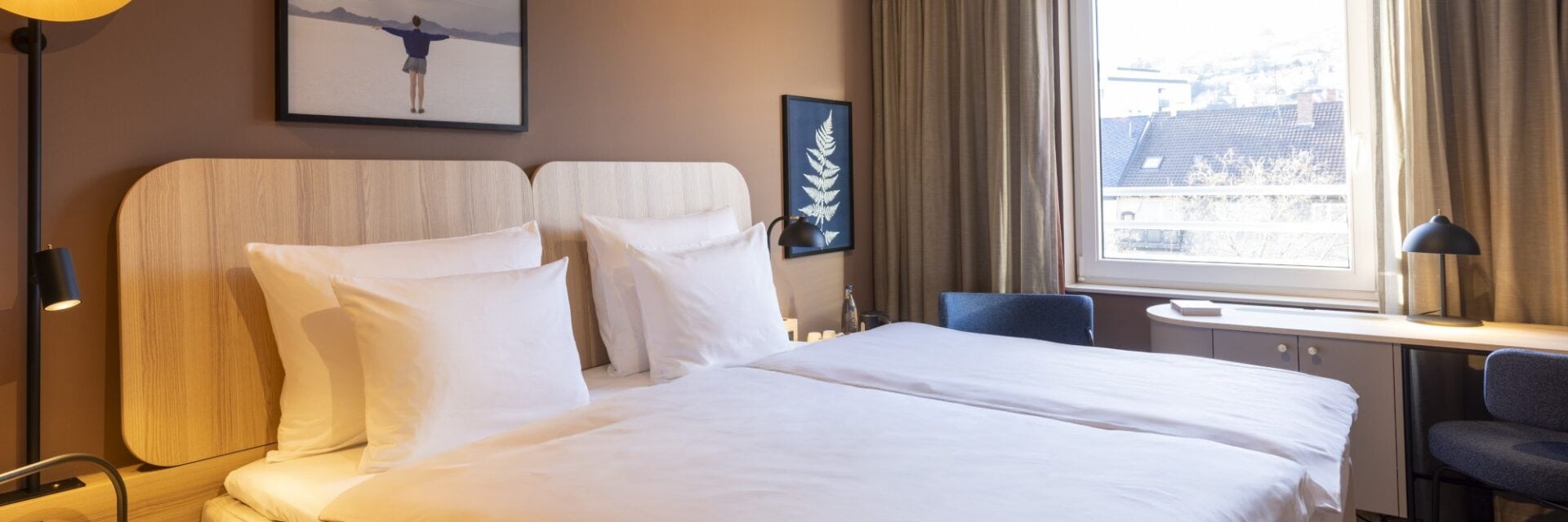 Ein Doppelbett steht in einem etwas dunkleren Zimmer, es ist auch das Fenster zu sehen.