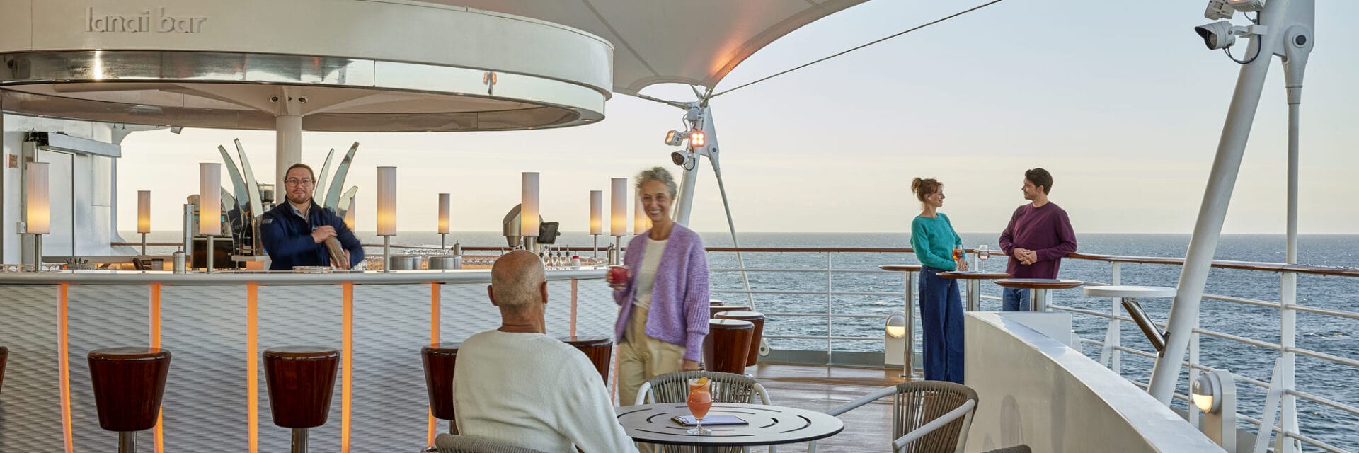 Offene weiße Bar am Heck eines Kreuzfahrtschiffes. Eine Handvoll Passagiere genießt den Blick aufs Meer.