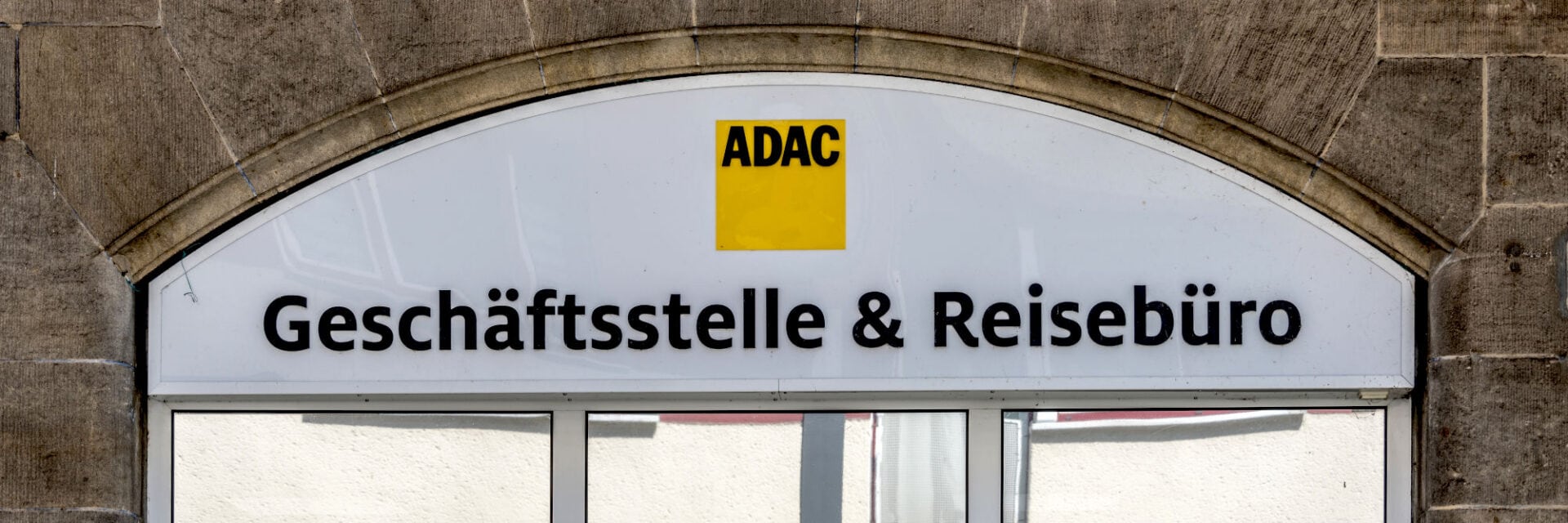 Das Bild zeigt den Eingangsbereich einer ADAC Geschäftsstelle und eines Reisebüros mit großem, weißem Schild und gelbem ADAC-Logo, umgeben von einem steinernen Rundbogen. Die Beschriftung „Geschäftsstelle & Reisebüro“ ist deutlich sichtbar.