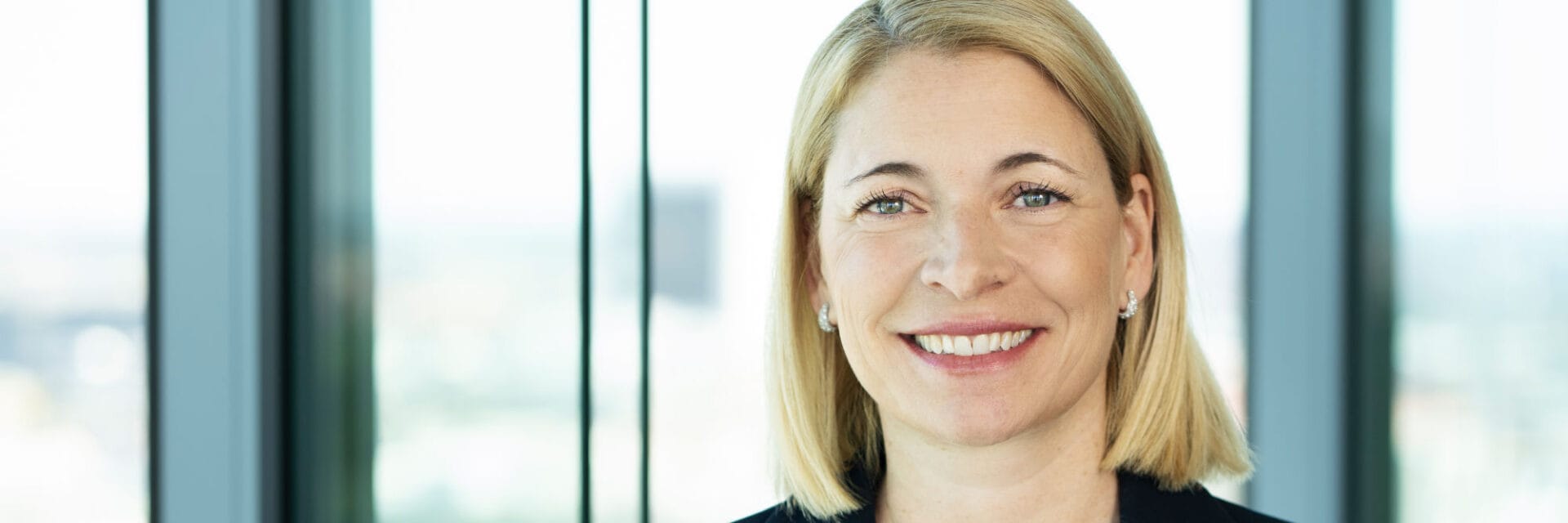 Porträt einer Frau mit blondem, schulterlangem Haar, die vor großen Glasfenstern in einem modernen Büro steht; das Bild eignet sich für Themen wie Business, Karriere und Management. Die neutrale Mimik und der unscharfe Hintergrund unterstreichen das professionelle Umfeld.