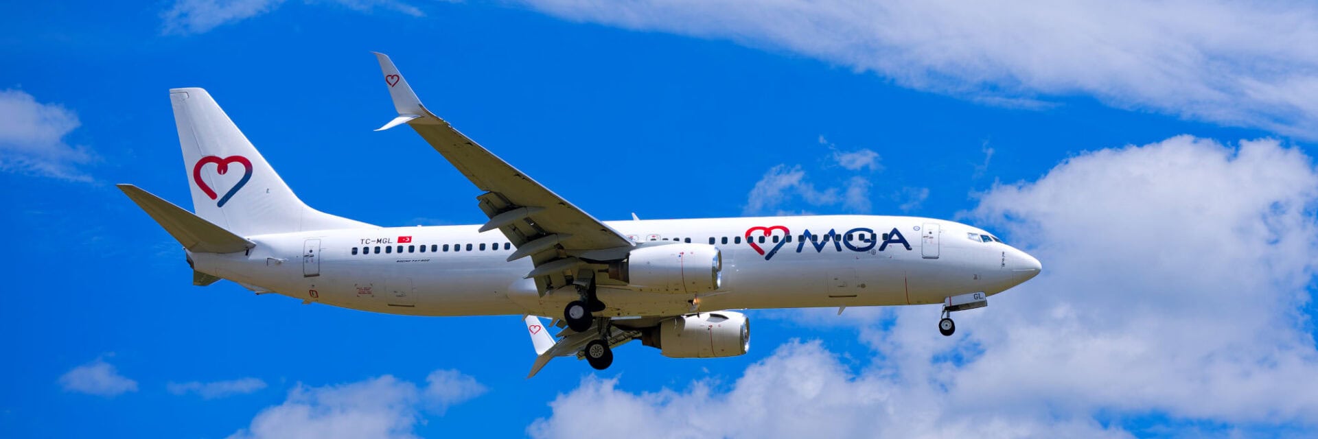 Ein weißes Passagierflugzeug der Airline Mavi Gök Aviation (MGA) fliegt bei Tageslicht vor einem strahlend blauen Himmel mit leichten Wolken. Das Flugzeug ist im Landeanflug, das Fahrwerk ist ausgefahren und das Firmenlogo mit Herzsymbol ist deutlich zu erkennen.